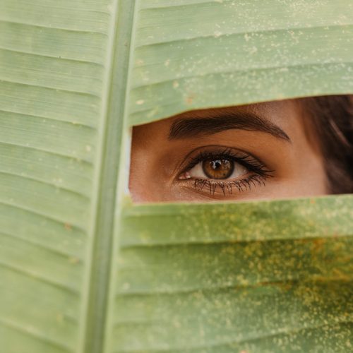 Dark-haired woman with green eyes looking into camera through hole in huge leaf